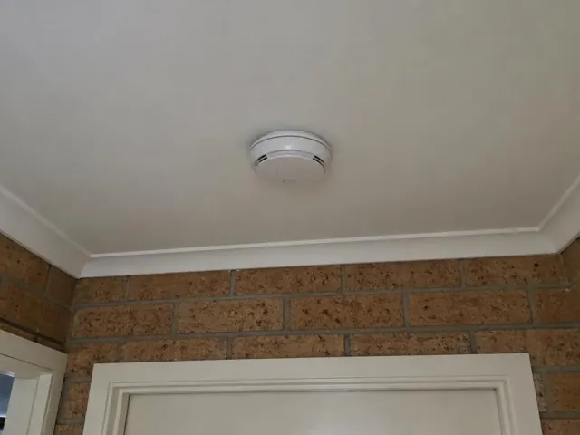 Ceiling-mounted smoke alarm on a plaster ceiling above a brick wall and doorway in Hamlyn Terrace